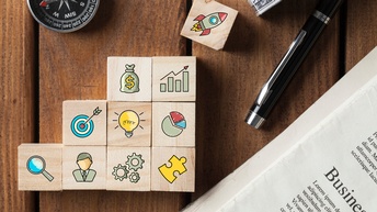 Auf einem braunen Holz-Untergrund liegt eine Zeitung, daneben ein Kugelschreiber und in der linken Bildhälfte ein Kompass. Mittig sind Holzwürfel zu Treppen gebaut. Darauf sind Symbole wie eine Rakete, Geldbeutel, Diagramme, eine Glühbirne, eine Lupe