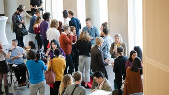 In einem großen Raum stehen in kleinen Gruppen verschiedene Leute, die miteinander reden. Manche von ihnen haben einen Ausweis um den Hals. Eine der Personen sitzt im Rollstuhl. Manche trinken einen Kaffee oder ein Wasser