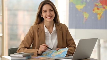 Eine lächelnde Person sitzt an einem Tisch vor einem aufgeklappten Notebook. In den Händen hält sie einen Zettel, auf dem eine Weltkarte abgebildet ist. An der Wand im Hintergrund verschwommen hängt noch eine Weltkarte.