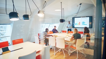 In einem Raum mit einer Glaswand auf einer Seite sitzen vier Personen um einen großen Tisch. Neben ihnen steht eine weitere Person, die neben einem großen Bildschirm steht. Auf dem Bildschirm sind Statistiken