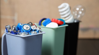 Drei Müllcontainer hintereinander stehend: Erster Container im Fokus gefüllt mit Batterien, dahinterstehende Mülleimer verschwommen gefüllt mit Plastikverschlüssen und Glühbirnen