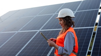 Person in oranger Schutzweste mit weißem Helm bedient Tablet, das sie in Händen hält, im Hintergrund schräg geneigte Solarpanele