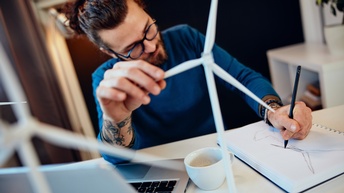 Person mit Brillen sitzt an Schreibtisch, skizziert Windrad auf Block und hält in einer Hand Modell eines Windrades, am Tisch aufgeklappter Laptop und Kaffeetasse