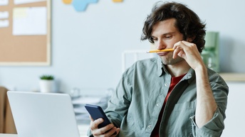 Eine Person sitzt an einem Tisch. Vor ihr ist ein aufgeklappter Laptop. Die Person hält ein Smartphone, auf das sie blickt. Mit der linken Hand hält sie einen Bleistift zwischen Nase und Oberlippe