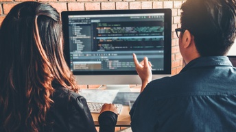 Zwei Personen in Rückenansicht sitzen vor Computerbildschirm, auf dem Quellcodes zu sehen sind