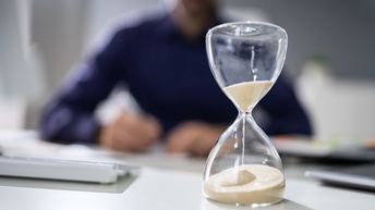 Sanduhr steht auf einem Schreibtisch, dahinter sitzt in Unschärfe eine Person, diverse Unterlagen liegen am Tisch