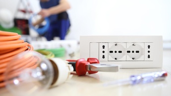 Tools für Arbeiten in der Elektronik wie Steckdose, Zange, Schraubenschlüssel und Kabel sowie Glühbirne, im Hintergrund zeigt sich eine Person, die in blauer Arbeitsbekleidung kniend Kabel in der Hand hält durchführt in der Unschärfe
