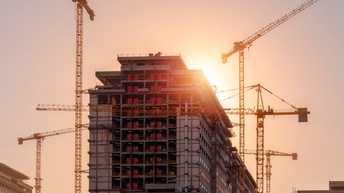 Ein mehrgeschossiges Gebäude befindet sich im Bau. Die Tragkonstruktion und mehrere Geschosse sind freigelegt. Rund um das Gebäude stehen mehrere Baukräne. Die Sonne befindet sich hinter dem oberen Gebäudebereich