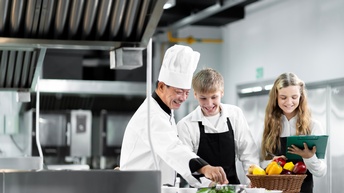 In einer Küche stehen zwei Jugendliche mit Schürzen und eine Person in weißer Kleidung und Kochhaube. Vor ihnen stehen Schüsseln und Teller. Daneben ist ein Korb mit Paprika.