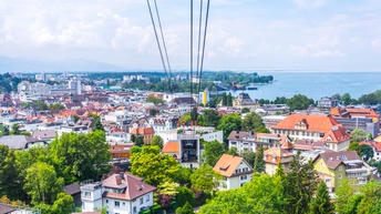 Sechs dunkle Seile gehen gespannt vom oberen Bildrand in die Bildmitte. Sie enden bei einem Gebäude, bei dem eine Seite komplett offen ist und man ist das Innere sieht. Um dieses Gebäude stehen weiter Gebäude. Dahinter ist ein Gewässer