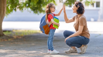 Seitliche Aufnahme eines Kindes mit Brille, Rucksack und Notizheften, die einer vor ihr hockenden Person gegenübersteht und mit der Person einschlägt. Kind und Person stehen in einem asphaltierten Innenhof neben einem grünen Baum