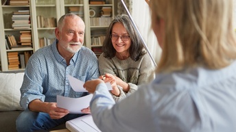 Im Bildvordergrund ist der Rücken einer Person mit blonden Haaren. Diese Person zeigt mit der linken Hand ein Blatt Papier zwei ihr gegenüber sitzenden älteren Personen, im Hintergrund Bücherregal.