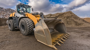 Ein Baufahrzeug mit einer großen Schaufel dran steht auf einem lehmigen Untergrund. Im Hintergrund sind mehrere Hügel aus verschiedenen Rohstoffen wie Steine oder Sand