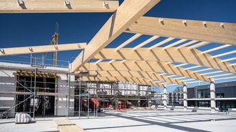 Auf einer Baustelle ist eine große, asphaltierte Fläche. Über der Fläche ist der Rahmen für ein Dach aus Holz gebaut. Auf dem Boden liegen Holzbalken. Im Hintergrund ist ein Rohbau aus Ziegeln, vor dem Baugerüste stehen