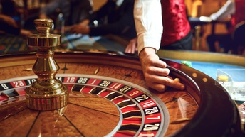 Nahaufnahme einer Holzscheibe, an die schwarze und rote Quadrate in einem Kreis angebracht sind. In jedem Quadrat steht eine Zahl. Die linke Hand einer Person hält eine kleine, helle Kugel an den erhöhten Rand des äußeren Kreises