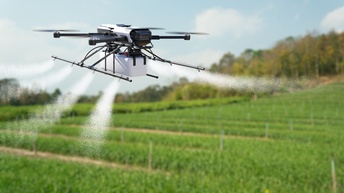 Eine kleine, weiße Drohne schwebt über einem grünen Feld. An dem unteren Part der Drohne sind lange Stangen, aus denen eine Flüssigkeit gesprüht wird.