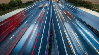 Foto einer Straße in Bewegungsunschärfe. Links und rechts von der Straße sind Felder. Auf der Straße fahren verschiedene Fahrzeuge wie Autos und Lastkraftwagen