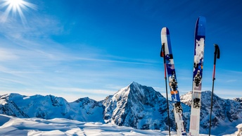 Zwei Ski stecken vertikal in einer Schneedecke. Links und rechts davon sind jeweils ein Skistock. Im Hintergrund sind schneebedeckte Berge. Der Himmel ist blau, man sieht in der linken Bildhälfte das Licht der Sonne. 