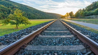 Schienengleise umgeben von grünen Feldern, Bäumen und Straße, im Hintergrund bewaldete Hügel und Sonne, die durch Laub eines Baumes blitzt