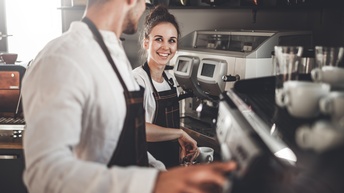 Person lächelt andere Person an, beide tragen Schürzen und stehen vor Kaffemaschine