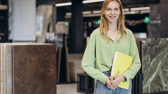 Lächelnde Person im Portrait mit gelber Mappe und Bleistift in Händen, im Hintergrund verschwommen Steinplattenmuster