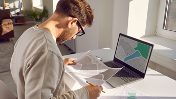 Bärtige Person mit Brillen in Seitenansicht sitzt an Arbeitsplatz vor aufgeklappten Laptop, am Bildschirm eine Landkarte mit nummerierten Parzellen ersichtlich, hält Ausdruck einer weiteren Karte mit nummerierten Parzellen in Händen und blickt darauf