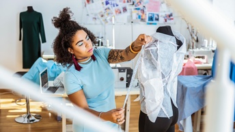 Person mit lockigem dunklen Haar steht bei einer Schneiderpuppe mit drapierten Stoffmuster und misst mit einem Maßband