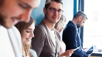 Mehrere Personen nebeneinander stehend, in verschiedene Smartdevices blickend, eine Person mit Brillen blickt in Kamerarichtung