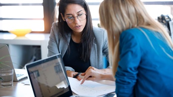 Zwei junge Frauen, eine dunkelhaarig mit Brille, die andere blond, sitzen bei einem Laptop und besprechen Unterlagen, die sie auf dem Tisch liegen haben.