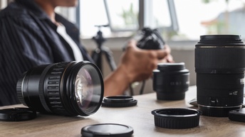Eine Person sitzt an einem Tisch und hält einen Fotoapparat. Auf dem Tisch sind verschiedene Kameraobjektive und -linsen