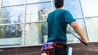 Rückenansicht einer Person mit Kappe, T-Shirt und einem Gürtel mit Fächern umgeschnallt, die vor einer Fensterfront steht und auf ein Fenster einen Wischer hält. Im Gürtel sind verschiedene Putzutensilien. Die Person