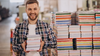 Porträt einer lachenden Person mit Bart und einem Stapel Büchern in beiden Händen. Hinter der Person ist ein hoher und breiter Stapel verschiedener, bunter Bücher