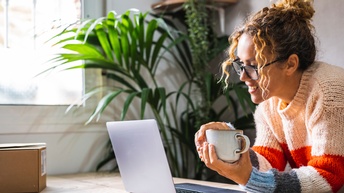Im rechten Bildrand beugt sich eine Person über einen hölzernen Untergrund und stützt sich mit beiden Unterarmen ab. In den Händen hält sie eine Tasse. Vor ihr steht ein aufgeklappter Laptop, auf den sie blickt.
