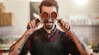 Frontale Aufnahme einer Person mit Schürze und Tattoos auf den Armen, die sich zwei runde Behälter mit Griffen vor die Augen hält. Die Behälter sind mit Kaffeepulver gefüllt. Im Hintergrund ist verschwommen eine Küche erkennbar