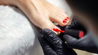 Nahaufnahmen der linken, rot lackierten Zehen einer Person in der linken Bildhälfte. In der rechten Bildhälfte sind die Hände einer anderen Person mit schwarzen Handschuhen, die die große Zehe hält und darauf roten Nagellack pinselt.