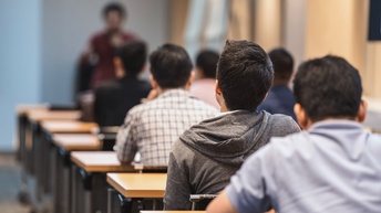 Rückenansicht von mehreren Personen, die hintereinander einzeln an einem Tisch sitzen. Ihnen gegenüber steht eine Person