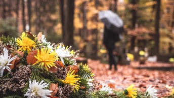 Links im Vordergrund liegt ein Blumenschmuck mit Tannenzweigen sowie weißen und gelben Blüten auf der Erde. Im Hintergrund ist die Rückenansicht einer Person, die zwischen Gräbern auf Laub steht und einen Regenschirm hält