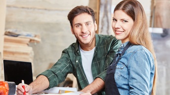 Zwei Personen stehen nebeneinander an einem Holztisch und blicken in die Kamera. Die rechte Person trägt eine Schürze. Die linke Person zeichnet mit einem Bleistift etwas auf ein großes Blatt Papier. Daneben liegt ein Maßband