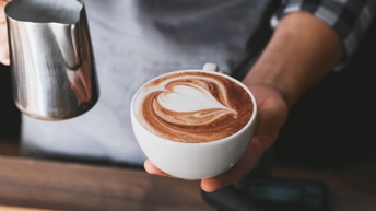 Detailansicht einer Person, die Kaffeetasse gefüllt mit Kaffee und Milchschaum in einer Hand hält, in anderer Hand Edelstahlmilchkanne