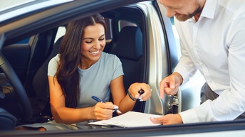 Eine Person sitzt in einem Auto auf dem Fahrersitz bei geöffneter Tür. Ihr gegenüber steht eine andere Person, die ihr ein Klemmbrett hinhält, auf dem ein Zettel ist. Die im Auto sitzende Person hält einen Stift mit dem sie unterschreibt 