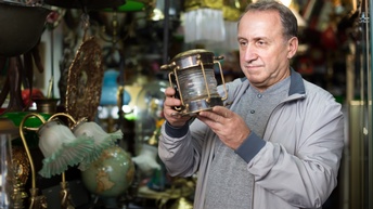 Person hält eine alte Lampe in der Hand und begutachtet sie, im Hintergrund befinden sich viele weitere Antiquitäten 