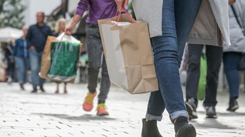 Im Vordergrund ist der Unterkörper einer Person, die mit einer Einkaufstasche in der rechten Hand durch eine Fußgängerzone geht. Im Hintergrund sind weitere Personen