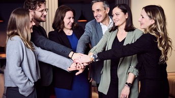 Eine Gruppe von Personen steht zusammen. Jede hat eine Hand ausgestreckt, die Personen führen die Hände in der Mitte zusammen und stapeln diese übereinander. 