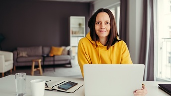 Person mit dunklen schulterlangen Haaren sitzt mit einem gelben Pullover an einem Schreibtisch mit einem Laptop, einem geöffneten Notizbuch sowie einem Smartphone, einer Tasse, einem Glas Wasser und einer Brille, im Hintergrund zeigt sich ein dunkler Wohnraum mit Sofa, Regalen sowie eine helle Fensterfront