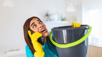 Eine Person mit gelben Handschuhen hält mit der linken Hand einen schwarzen Kübel und mit der rechten Hand ein Telefon an ihr rechtes Ohr. In den Kübel fallen von oben Wassertropfen. Die Person blickt auf die Wassertropfen