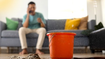 Im Vordergrund steht auf einem Holzboden in einem Wohnraum ein roter Kübel, in den von oben Wasser tropft. Neben dem Einer ist ein Lappen. Im Hintergrund sitzt eine Person auf einem Sofa und telefoniert