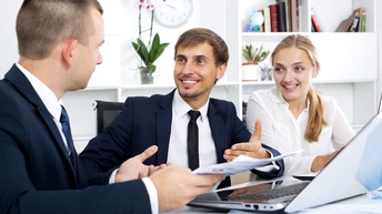 Hier sind 3 Beamte zu sehen (2 Männer, 1 Frau). Sie diskutieren miteinander über den Inhalt eines Blattes und eines Laptops. Sie vermitteln eine locke und gemütliche Arbeitsatmosphäre. Im Hintergrund sieht man Kästen mit Büchern und Pflanzen.