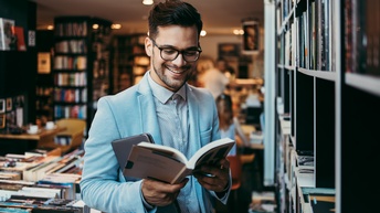 Lächelnde Person mit Brillen steht in Buchhandlung und blickt in aufgeschlagenes Buch
