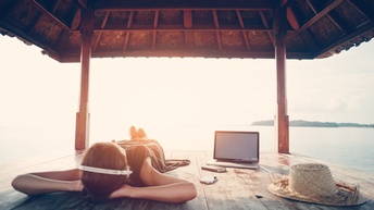 Eine Person liegt mit Laptop und Kopfhörern im Schatten eines Unterstands an einem Gewässer und hört Musik, während Sonnenlicht blendend einfällt. Neben ihr liegt ein Strohhut.