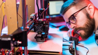 Person mit Brille, Bart und kurzen dunklen Haaren blickt auf einen 3D-Drucker, der gerade ein Material verarbeitet